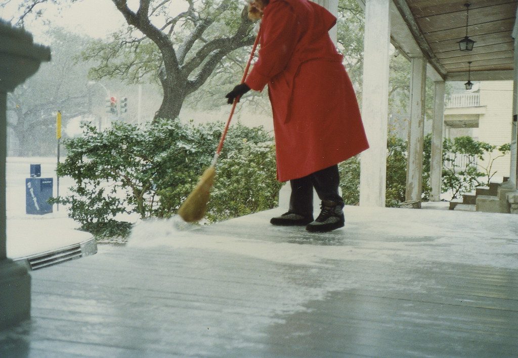A person in a red coat sweeps snow off a porch with a broom